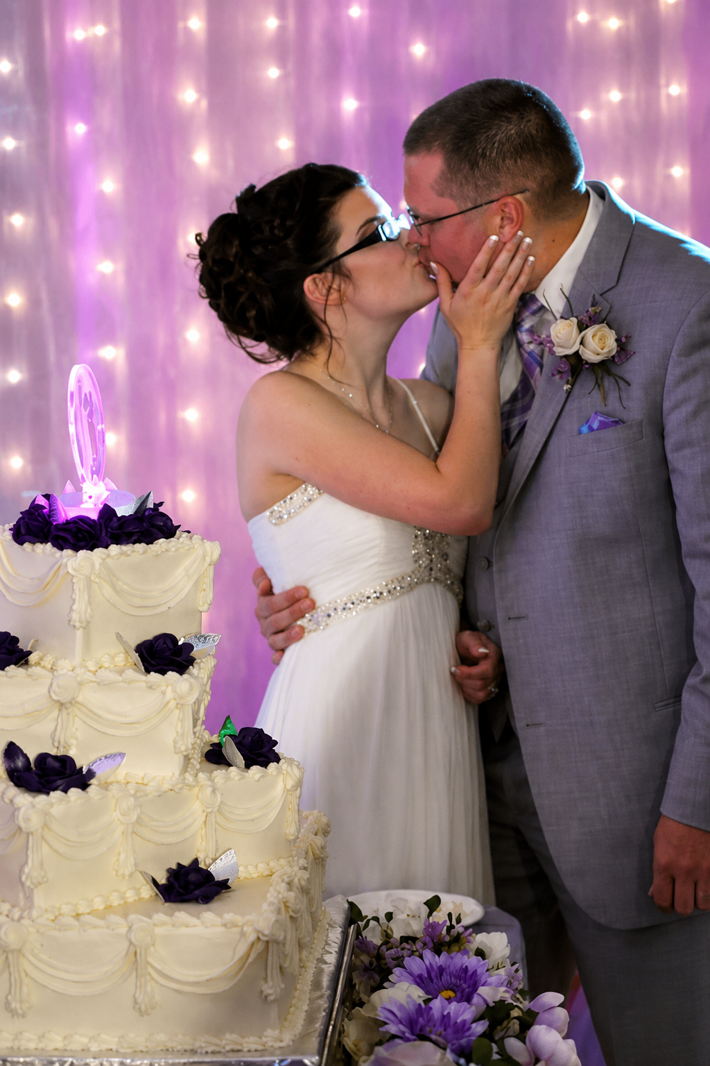 Bride and groom sharing a kiss beside their wedding cake with purple fairy light backdrop