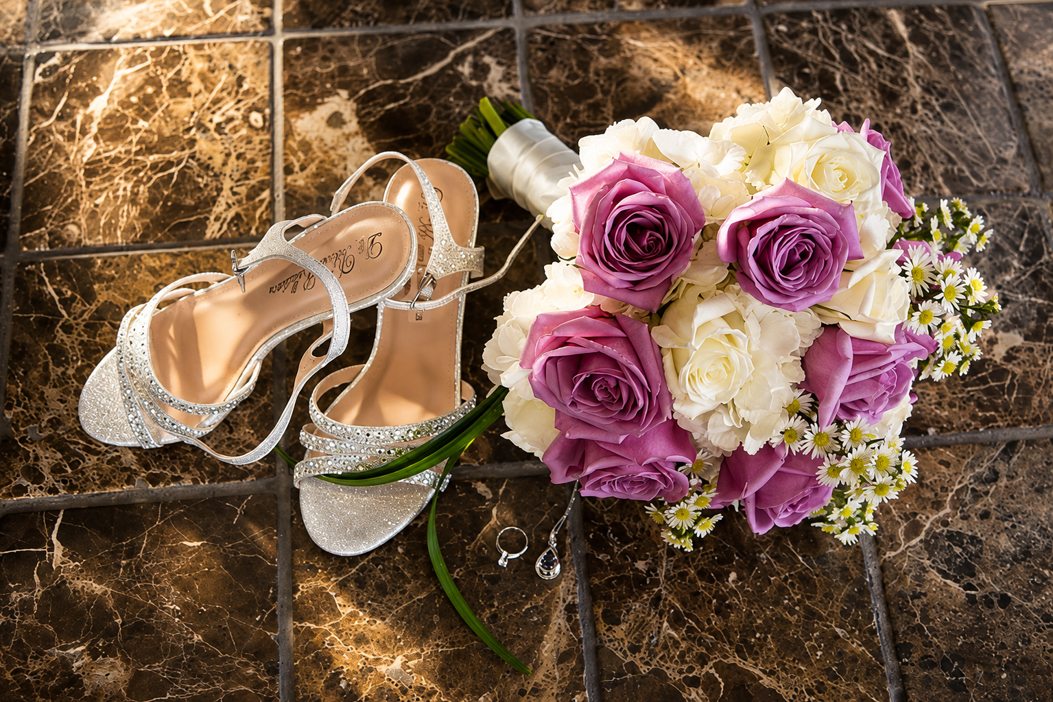 Bridal flat lay with silver rhinestone heels, purple and white rose bouquet, ring and earrings on marble tile