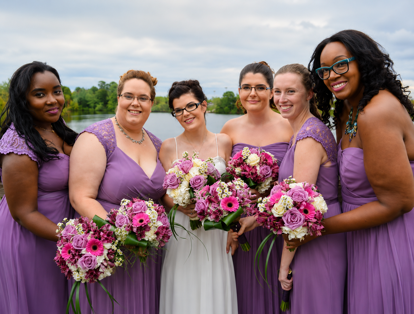 Bride and bridesmaids in purple gowns holding pink and lavender bouquets by a lakeside